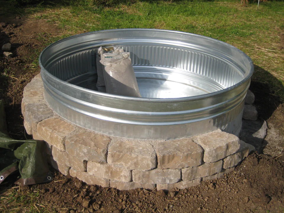 One galvanized stock tank dropped onto a carefully leveled circle of yard with a dry-stacked stone-block surround. The ring hides the bare tank wall for a better aesthetic.