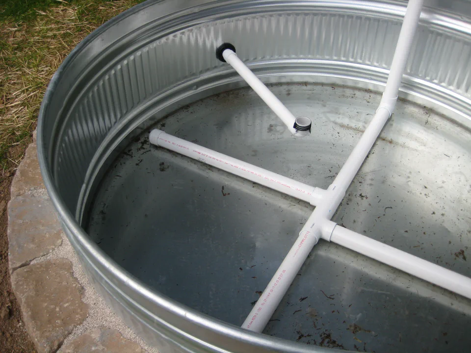 Inside the floor: a PVC distribution manifold. Water enters at the bottom and spreads across the whole footprint instead of pooling in one spot. The vertical pipe is the fill riser; the threaded port feeds out through the wall as an overflow.