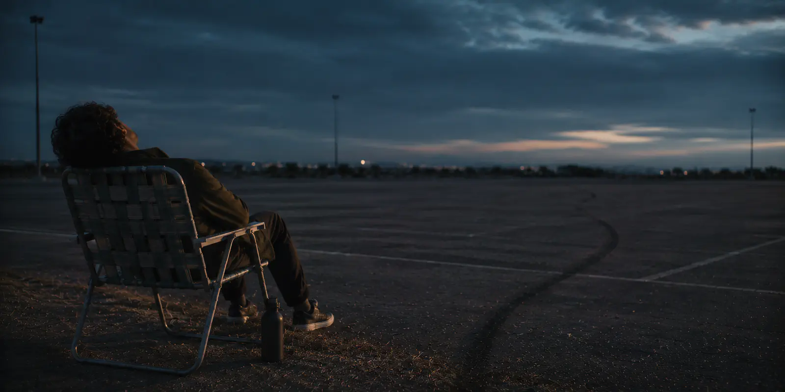 A man slumped asleep in a folding lawn chair beside a single tire track curving away across an empty lot at dusk.